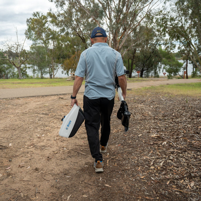 A man is walking on a dirt path carrying a Bixpy electric motor battery pack and outboard motor assembly towards the water.