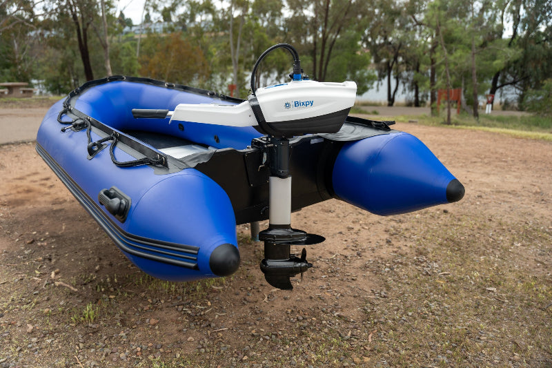 White Bixpy electric outboard motor mounted on the stern of a blue inflatable dinghy resting on land in an outdoor setting.