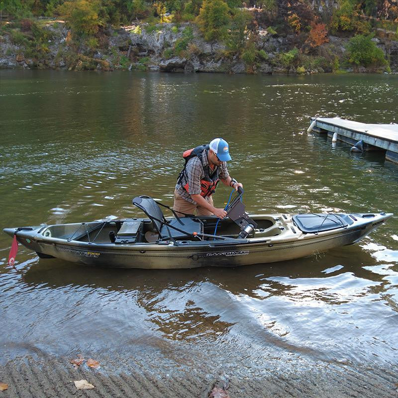 Man in a camouflage kayak in shallow water, installing a Bixpy motor and adapter into the kayak's pedal drive well next to a wooden dock.