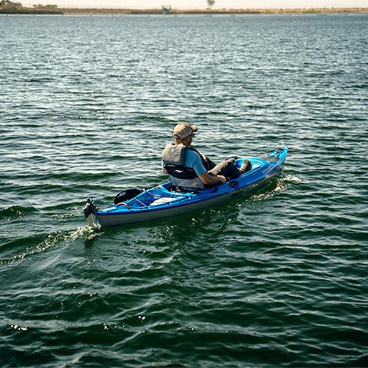 Man in a blue kayak propelled by a Bixpy Jet electric motor, leaving a wake on rippled water with a distant shoreline.
