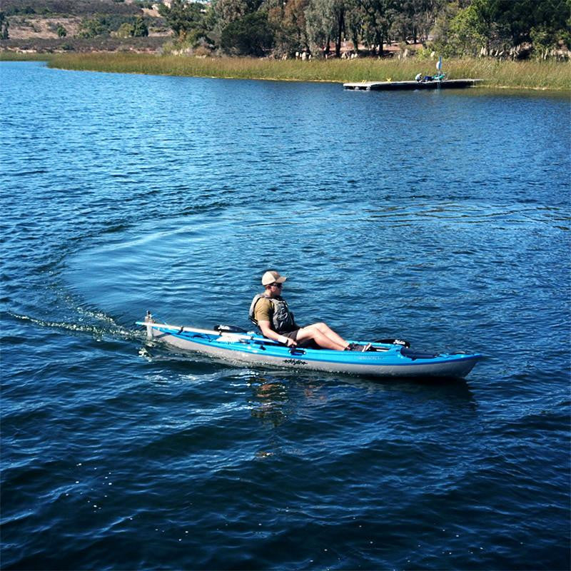 Man in a blue kayak propelled by a Bixpy Jet electric motor, leaving a wake on rippled water with a distant shoreline and dock.