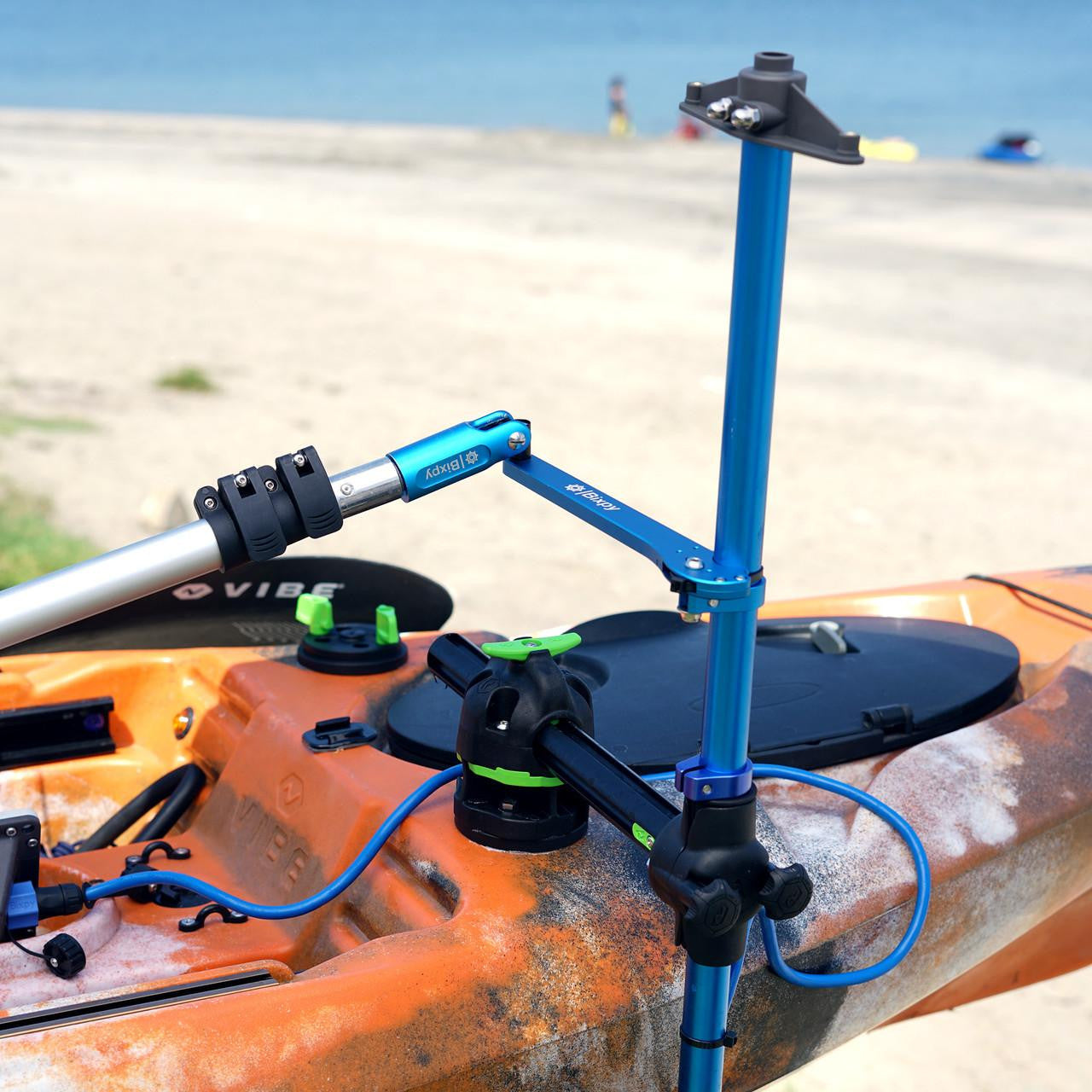 Close-up of a blue Bixpy pole mount adapter clamped to an orange camouflage Vibe kayak on a sandy beach, showing adjustable joints and paddle mount.