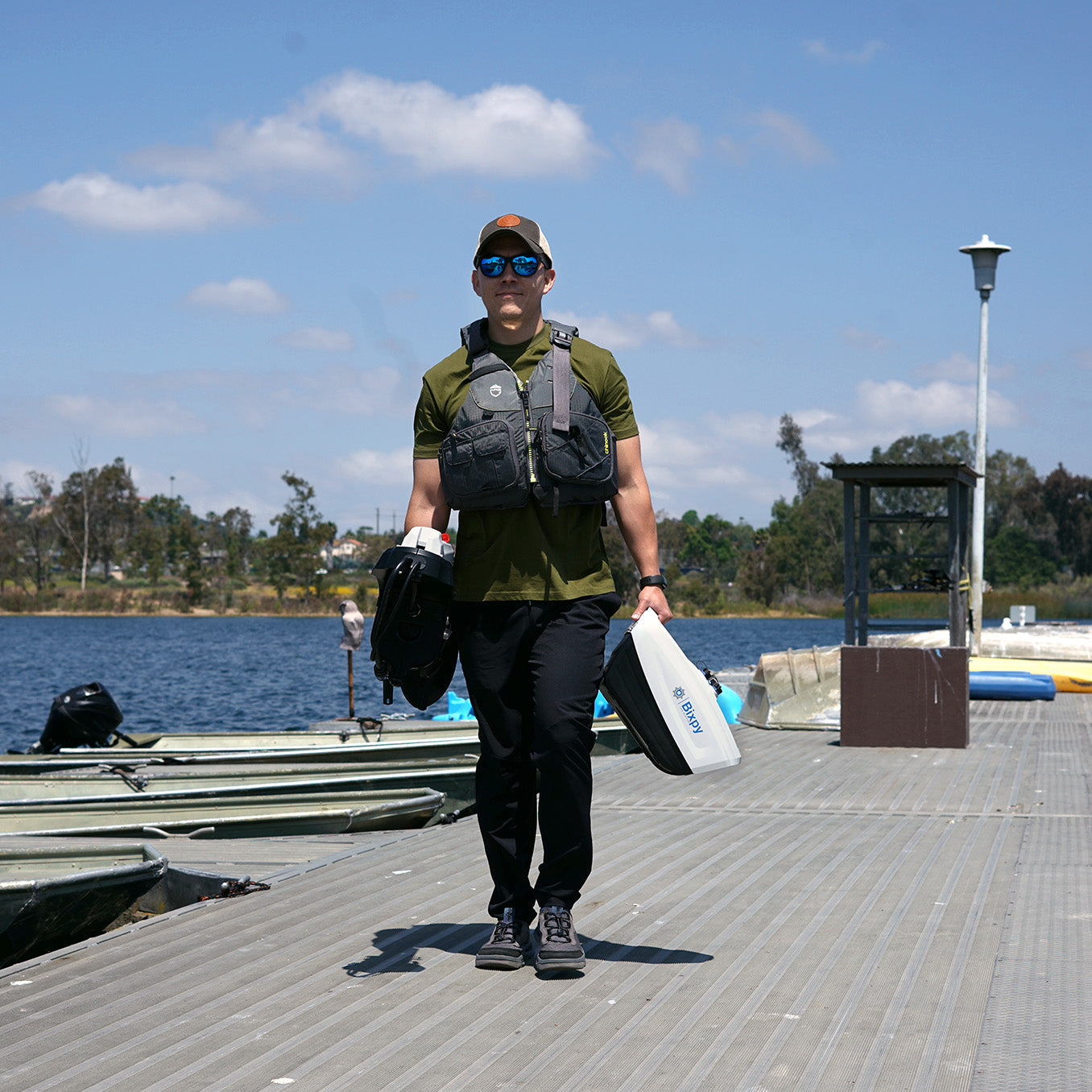 A man is walking on a dirt path carrying a Bixpy electric motor battery pack and outboard motor assembly towards the water.