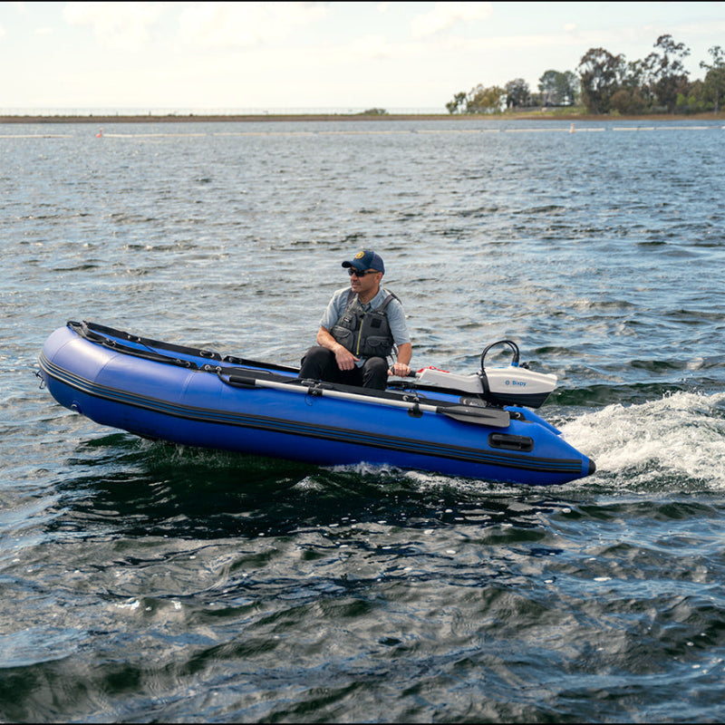 Man in a blue inflatable dinghy propelled by a white Bixpy electric outboard motor creating a large wake on rippled water.