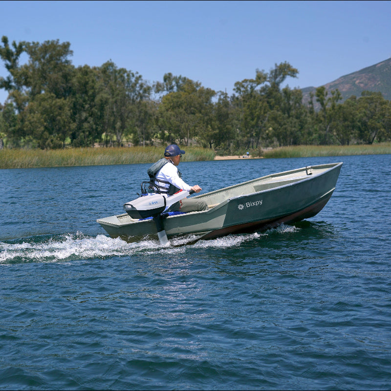 Man propelling a small light green jon boat with a white Bixpy electric outboard motor, creating a wake on open water with trees and hills in background.