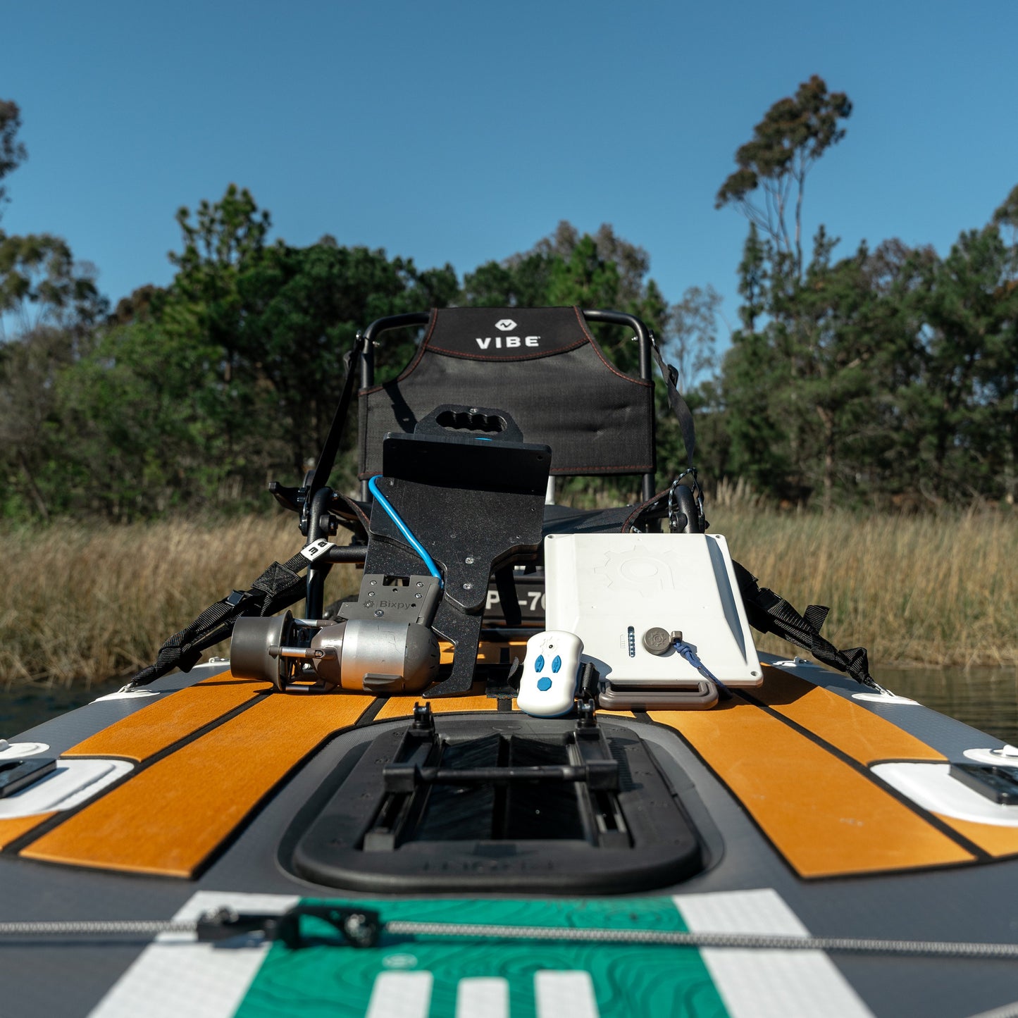 Front view of a multi-colored Vibe kayak with a Bixpy Jet electric motor, battery, and remote control, fully rigged with fishing rods on water near a treed shoreline.