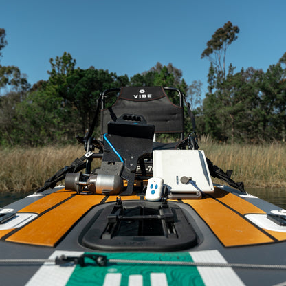 Front view of a multi-colored Vibe kayak with a Bixpy Jet electric motor, battery, and remote control, fully rigged with fishing rods on water near a treed shoreline.