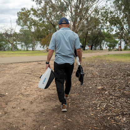 A man is walking on a dirt path carrying a Bixpy electric motor battery pack and outboard motor assembly towards the water.