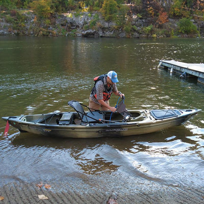 Man in a camouflage kayak in shallow water, installing a Bixpy motor and adapter into the kayak's pedal drive well next to a wooden dock.