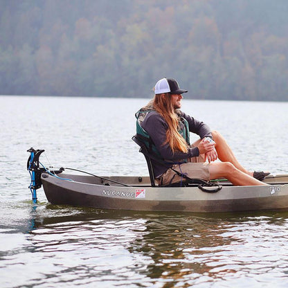 Man in a grey Nucanoe kayak propelled by a Bixpy Jet electric motor mounted on a blue vertical pole, on calm water with a forested shoreline.