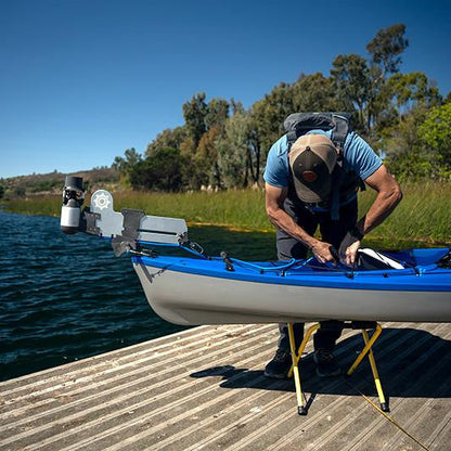 Man adjusting a Bixpy Jet electric motor mounted on a light gray Bixpy Versa Rudder on a blue and white kayak, resting on yellow stands on a wooden dock.