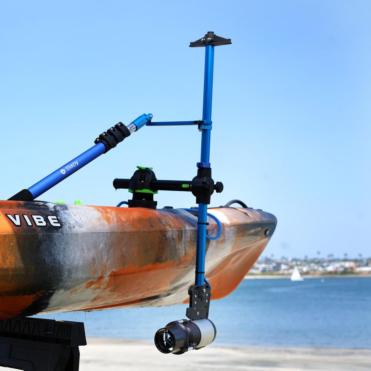 Orange and blue Vibe kayak with a Bixpy Jet electric motor mounted via a blue pole mount adapter, positioned above calm water with distant land and a sailboat.